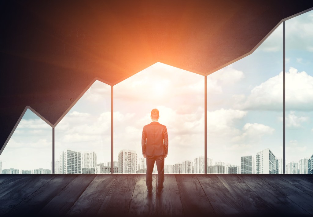 Businessman standing on the floor back to us looking through the big window with cityscape and sunset behind it.