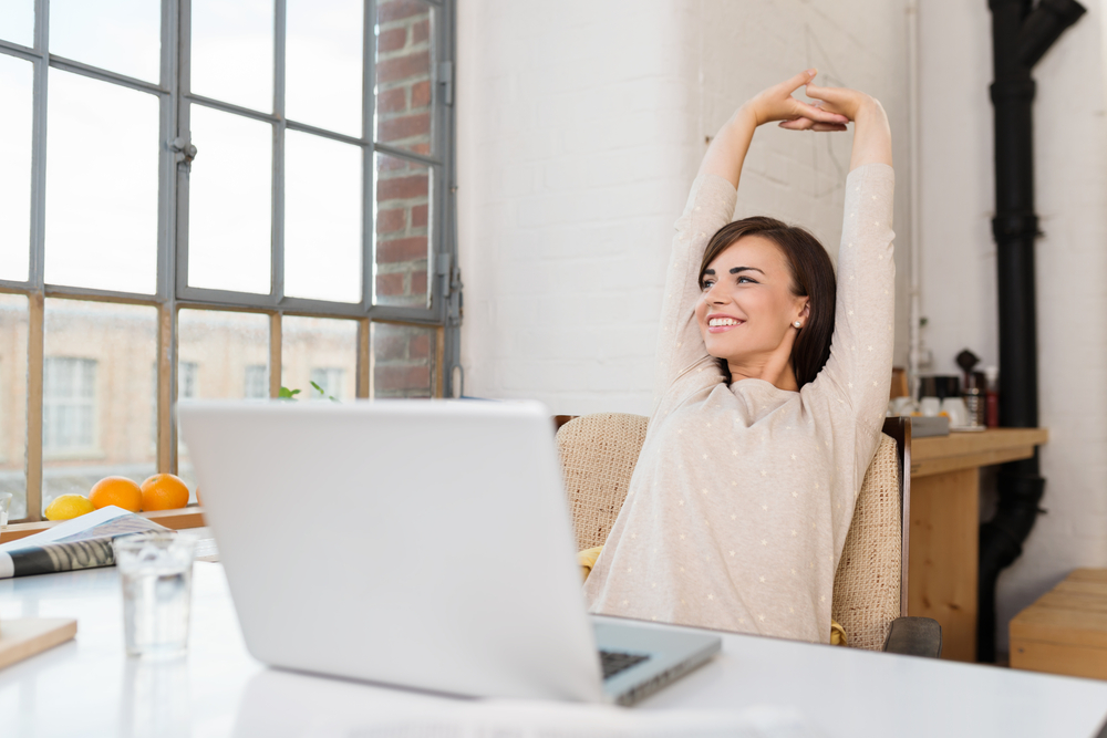 Happy-relaxed-young-woman-sitting-in-her-kitchen-with-a-laptop-in-front-of-her-stretching-her-arms-above-her-head-and-looking-out-of-the-window-with-a-smile