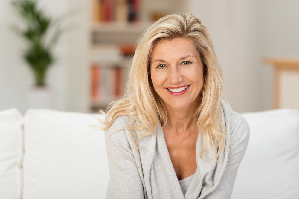 Lovely-middle-aged-blond-woman-with-a-beaming-smile-sitting-on-a-sofa-at-home-looking-at-the-camera
