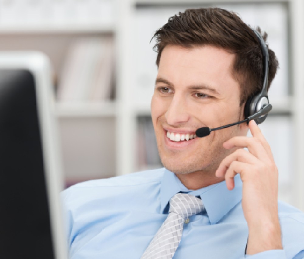 Smiling-friendly-handsome-young-male-call-centre-operator-or-client-services-personnel-beaming-as-he-listens-to-a-call-and-checks-information-on-his-computer-monitor