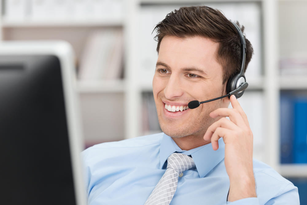 Smiling-friendly-handsome-young-male-call-centre-operator-or-client-services-personnel-beaming-as-he-listens-to-a-call-and-checks-information-on-his-computer-monitor