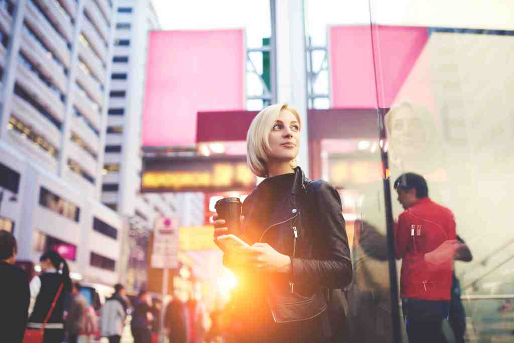 Mindful attractive blonde with trendy short haircut waiting for guide to looked at Hong Kong city planning sightseeing tour in morning drinking coffee to keep warm standing outdoors using free wifi
