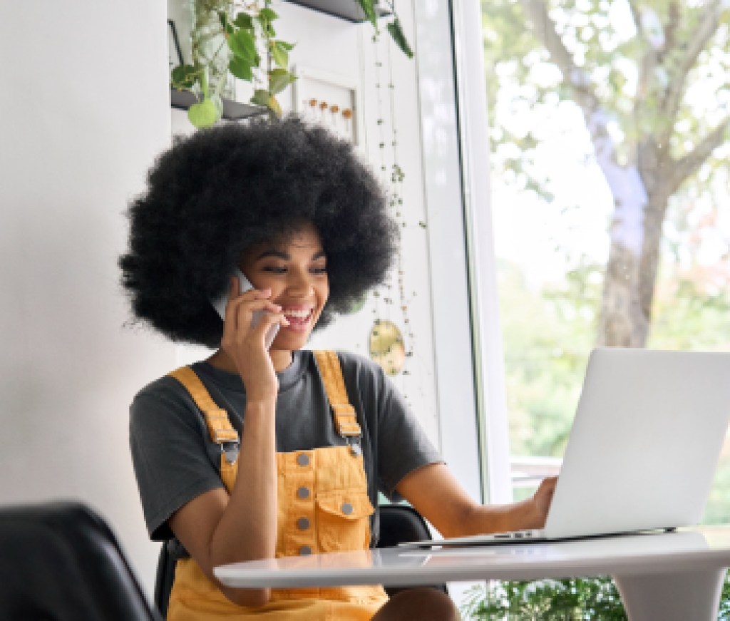 Young happy black college student speaking on phone using laptop in cafe.