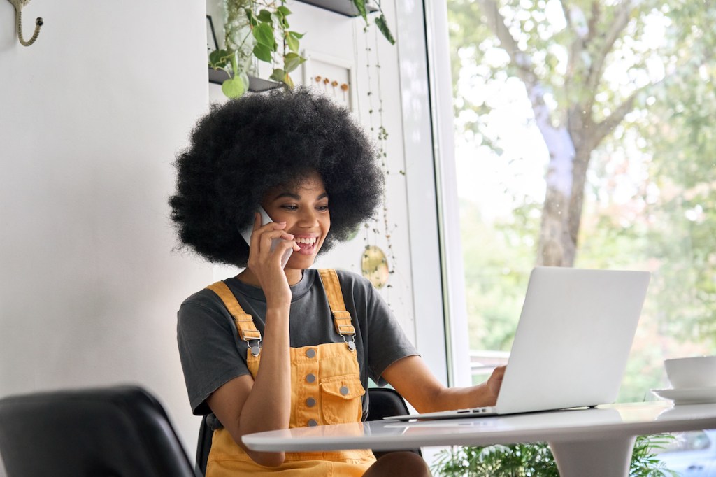 Young happy black college student speaking on phone using laptop in cafe.