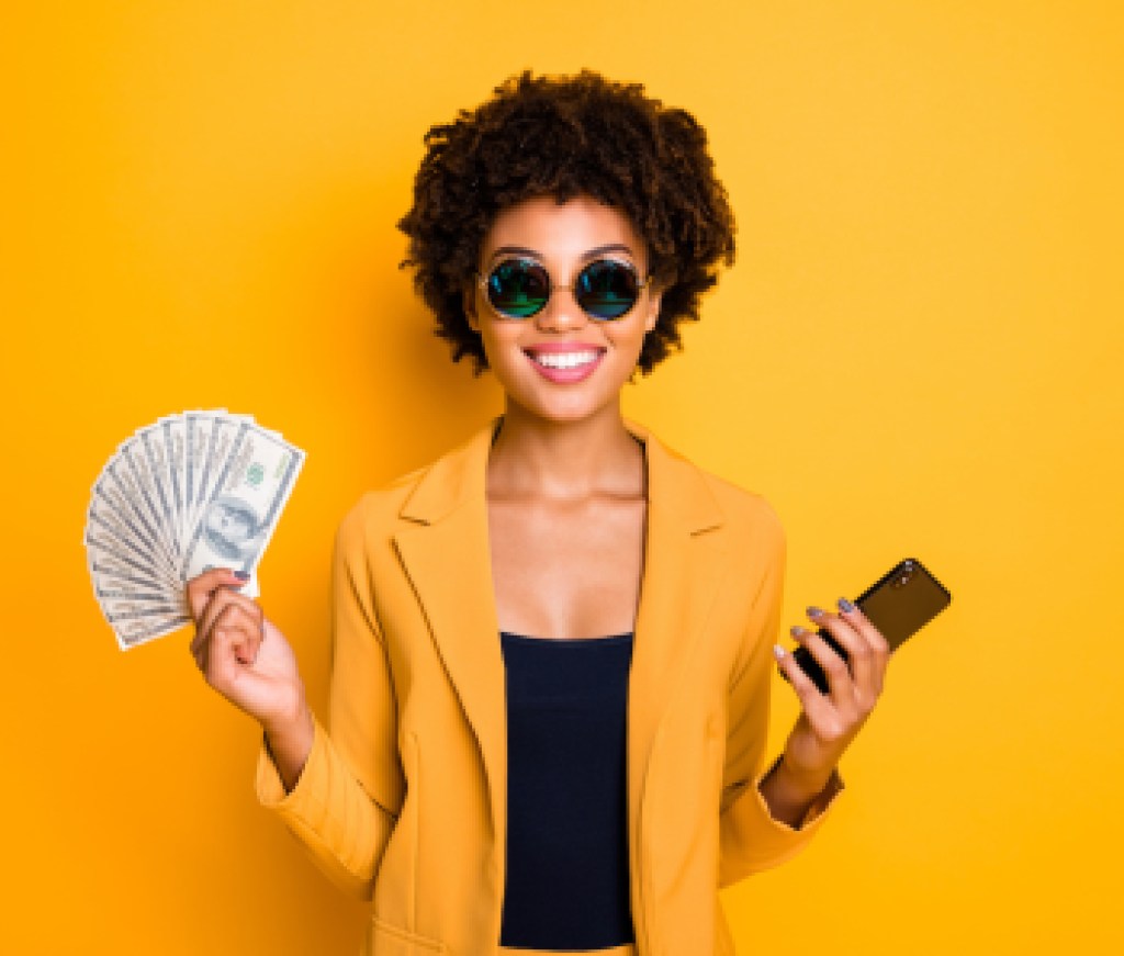 Portrait of positive cheerful afro american lady freelancer get cashback salary hold money fan use smartphone buy purchase pay online wear style blazer suit isolated over yellow color background
