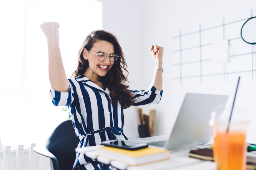 Excited young woman sitting at table with laptop and celebrating success