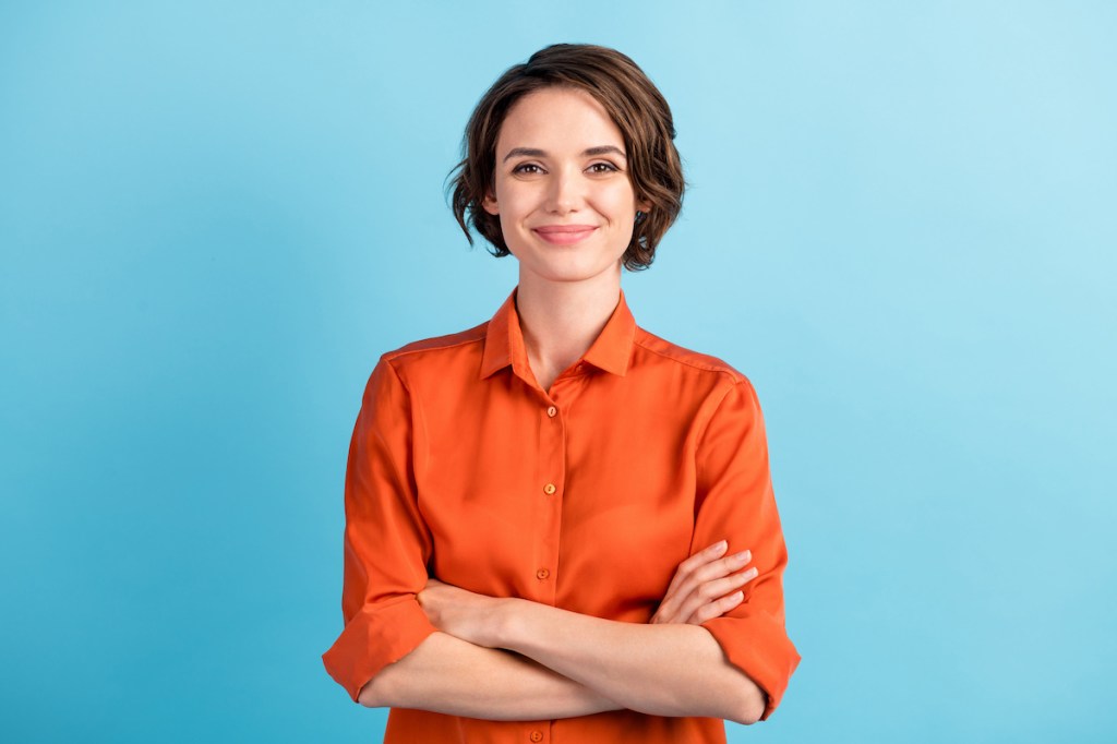 Photo of attractive charming lady cute bobbed hairdo arms crossed self-confident person worker friendly smile good mood wear orange office shirt isolated blue color background